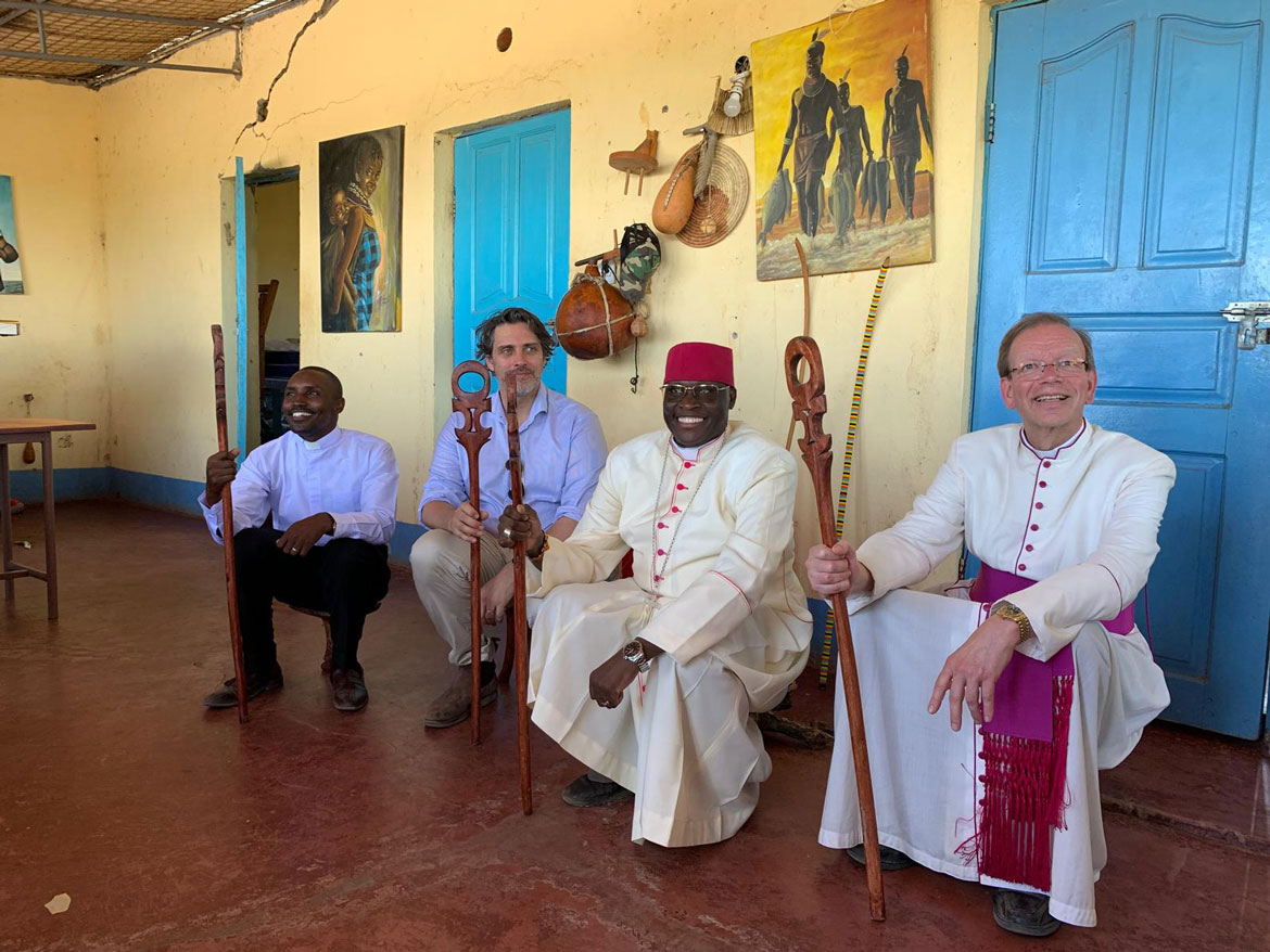 Ein kenianischer Geistlicher, Prinz Ludwig, der örtliche Bischof und Monsignore Wolfgang Huber sitzen vor einem Gebäude auf niedrigen Holzschemeln und halten Holzstäbe mit kunstvoll gestalteten Griffen in der Hand.
