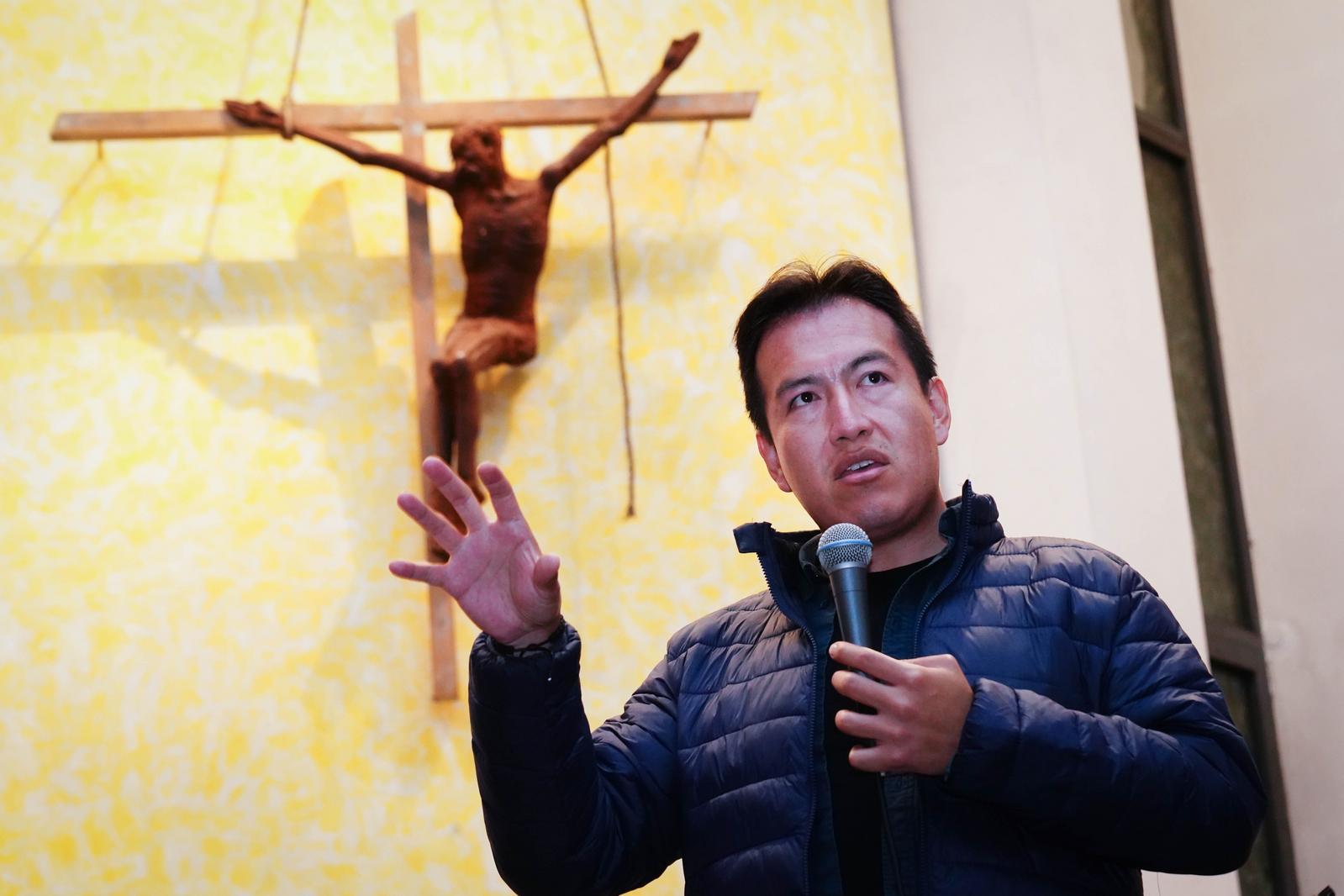 Pater Miguel Condo Soto hält ein Mikrophon in der Hand und spricht. Im Hintergrund ist ein großes Kruzifix zu sehen.