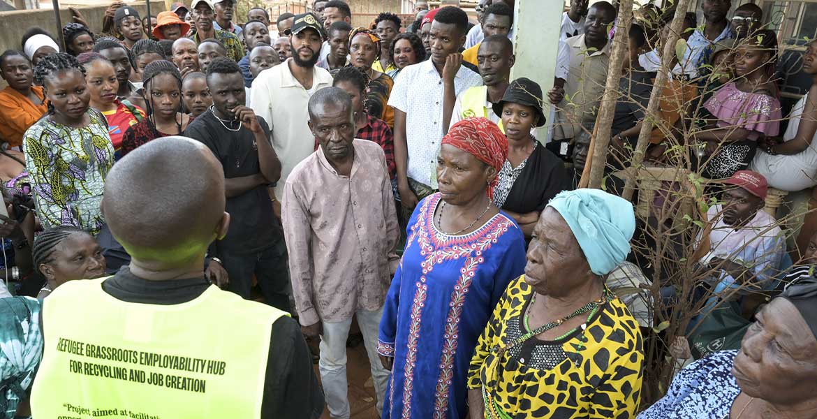 Ein Ordner steht (mit dem Rücken zur Kamera) in einer neongelben Weste in Kampala/Uganda vor einer Gruppe von geflüchteten Menschen aus dem Kongo. Foto: Jörg Böthling/missio München