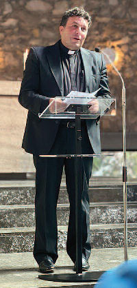 Maroniten-Pater Gaby Geagea steht vor einem Mikrophon und spricht. Hinter ihm führen Stufen zu einem Altar.