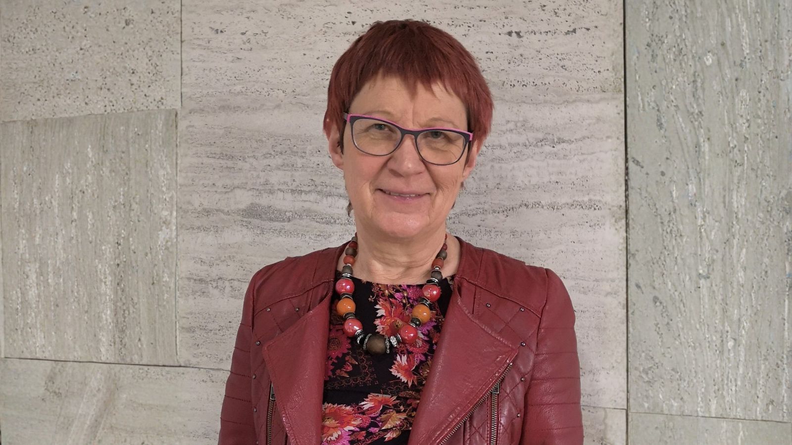 Porträtbild von Marita Krauss, die vor einer Wand aus Stein steht. rechts und links wurde der Hintergrund mit KI erweitert, um das erforderliche Bildformat zu erhalten.