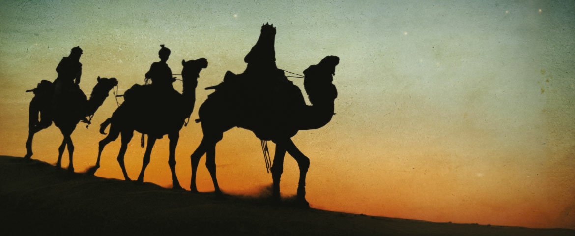 Schattenriss der heiligen drei Könige, die hintereinander auf Kamelen durch die Wüste reiten, im Hintergrund färbt die bereits untergegangene Sonne den Horizont in orangenes Licht