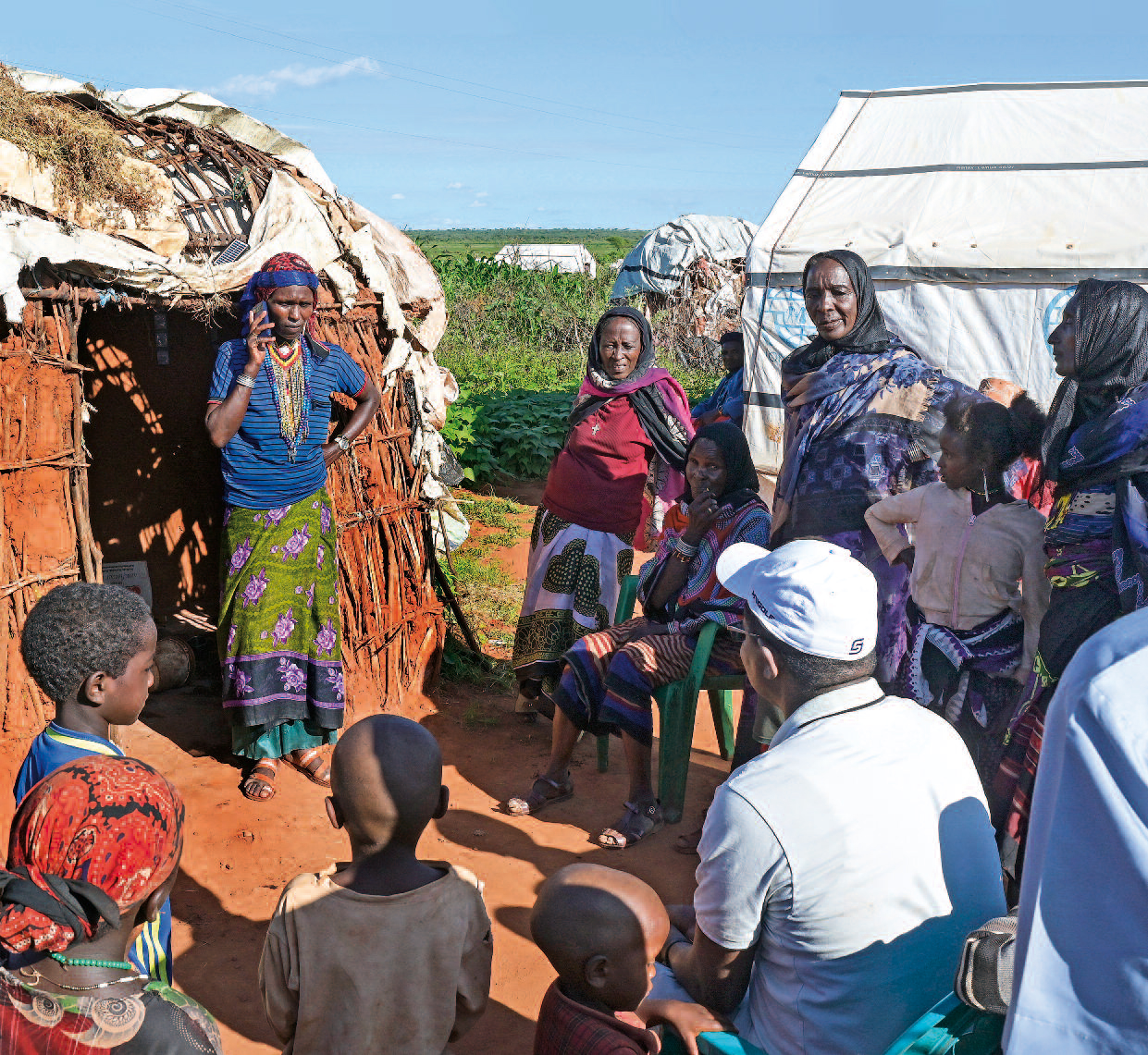 Eine Äthiopierin mit T-Shirt, einem langen, gewickelten Rock steht vor der Eingangstür zu einer Hütte und telefoniert. Um sie herum stehnd und sitzend Bewohnerinnen und Bewohner des Camps, darunter auch Kinder.