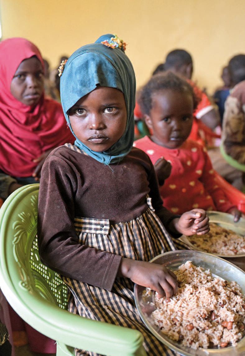 Ein kleines Mädchen mit Kopftuch sitzt auf einem Plastikstuhl und hält eine Metallschüssel mit reis und ein paar Bohnen auf dem Schoß, in die sie mit der rechten Hand greift. Um sie herum weitere Kinder mit verschrecktem Blick in ähnlicher Pose.