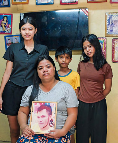 Roda sitzt mit dem Bild ihres Mannes in den Händen auf einem Stuhl. Drei Kinder stehen dahinter.
