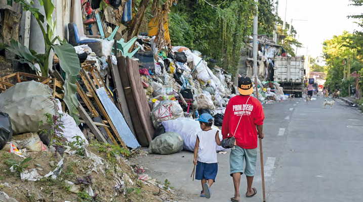 Ein Mann geht mit einem Kind an der Hand durch eine Straße. Am linken Wegrand türmen sich Müll und alter Hausrat.