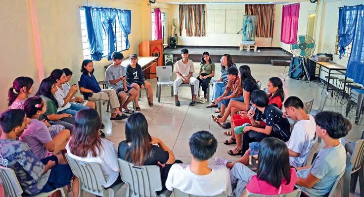 Eine große Gruppe von Jugendlichen sitzen auf Plastikstühlen in einem Raum. unter ihnen Carol Daria von Projekt "Solidarity with Orphans and Widows".