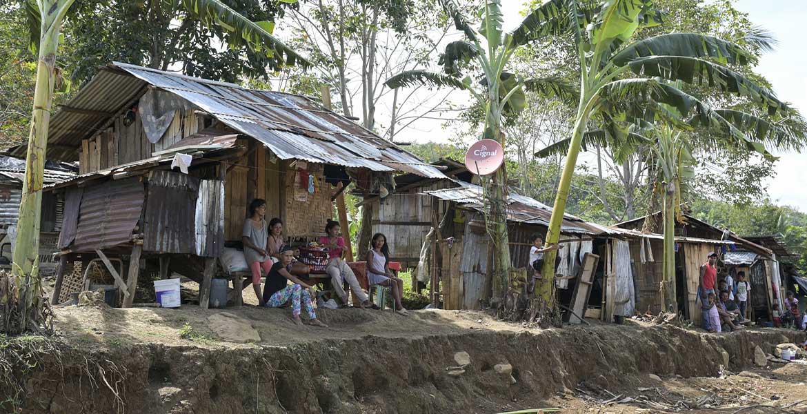 Menschen sitzen auf der philippinischen Insel Mindanao vor ihren ärmlichen mit Wellblech bedeckten Behausungen. Dazwischen wachsen Palmen.