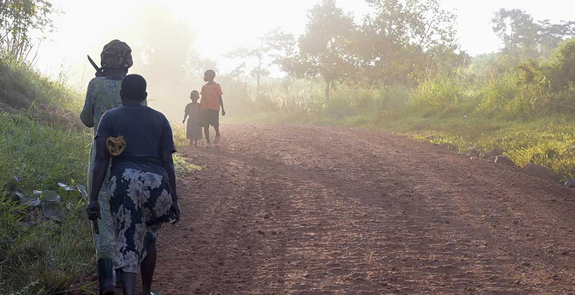 Auf der Insel Buvuma in Uganda laufen zwei Frauen auf einem am Rande eines breiten Feldwegs einen Berg hinauf. Sie sind von hinten zu sehen. Weiter vorne ist eine weitere Frau und ein Kind im nebeligen Sonnenlicht zu erkennen.