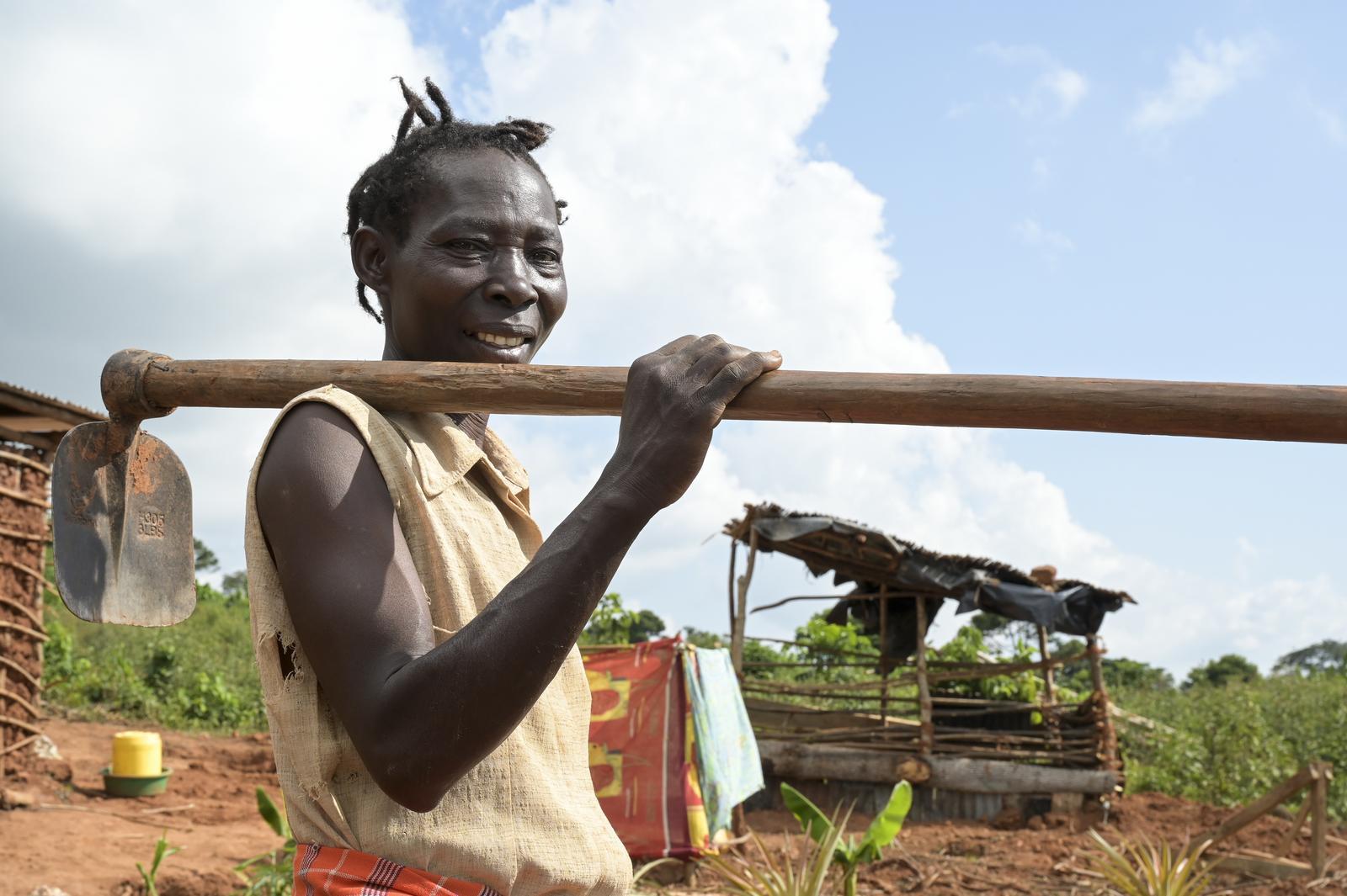 Eine Frau, Rose Nanteza, trägt auf der Schulter einen Spaten. Hinter ihr sind ein aus Holz gezimmerter Verschlag und spärliche Felder zu sehen. 