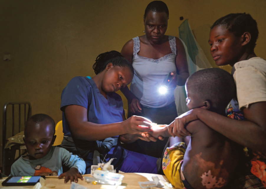 In einem dunklen Zimmer versucht eine Krankenschwester ein Kind zu behandeln, das neben seiner Mutter sitzt, die den Arm um ihn legt. Eine andere Frau leuchtet mit einer Taschenlampe den Arm des Jungen an. Ein kleines Kind sitzt daneben und blickt gebannt auf ein Tablet.