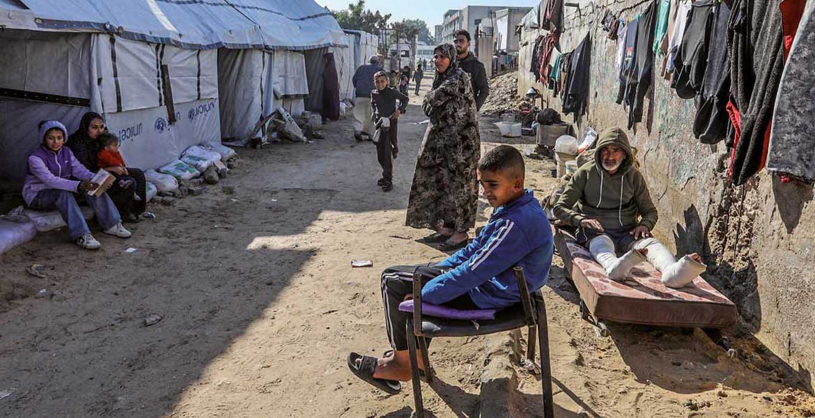 Geflüchtete Palästinenser aus dem Gazastreifen sitzen und stehen auf einer Lehmstraße. Links sind Zelte zu sehen, rechts aufgehängte Wäsche an einer Mauer.
