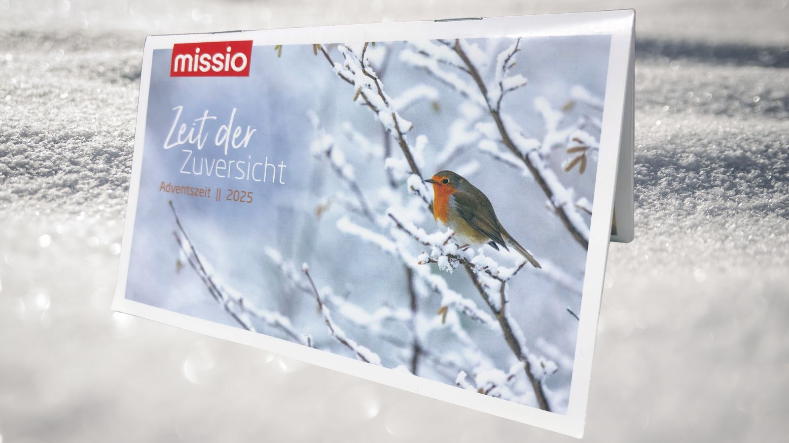 Der missio-Adventskalender 2025 steht im Schnee. Er zeigt ein Rotkehlchen auf verschneiten Ästen und trägt den Schriftzug Zeit der Zuversicht
