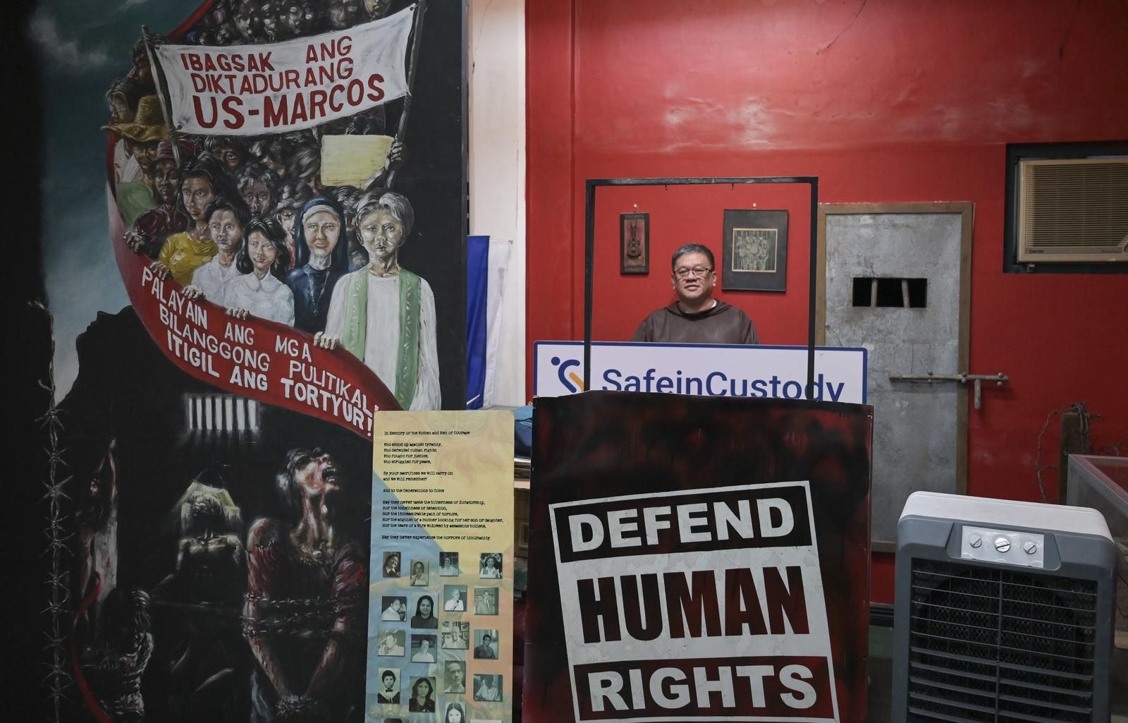 Father Toots steht in einem rot gestrichenen Raum neben einem Wandgemälde zum Andenken an die Widerstandskämpfer unter der Marcos Diktatur.