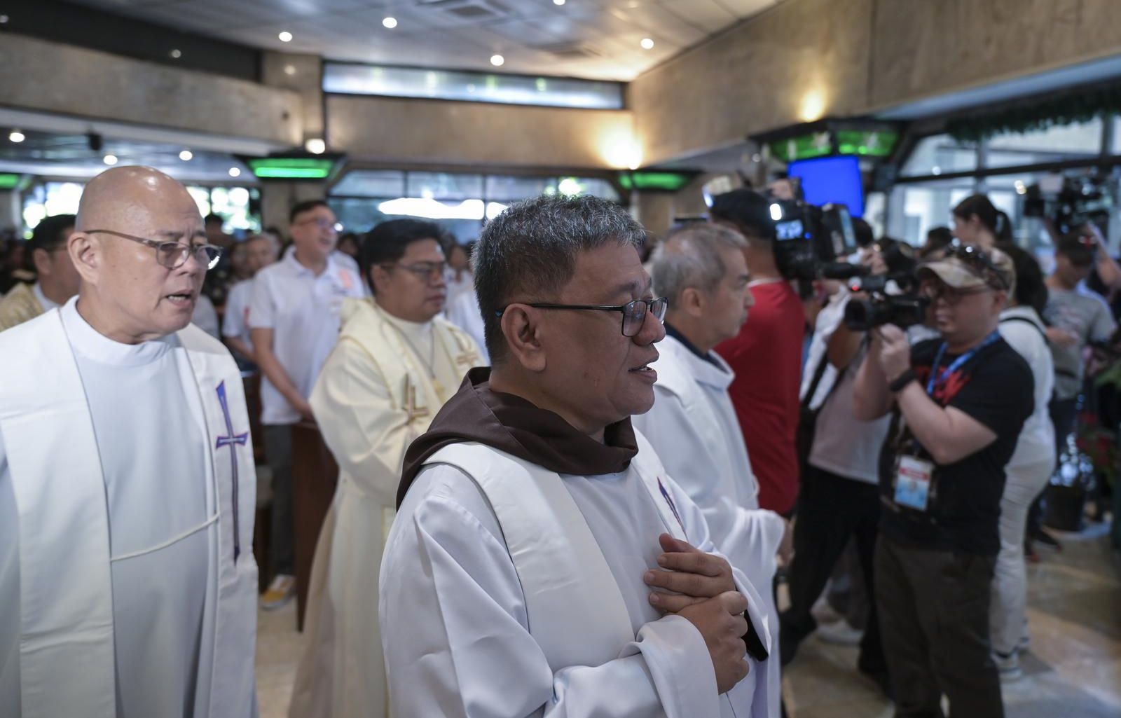 Father Toots trägt ein Messgewand. Er steht zusammen mit anderen Ordensleuten in einer großen, gut gefüllten Halle. Ein Kameramann filmt die Szene.