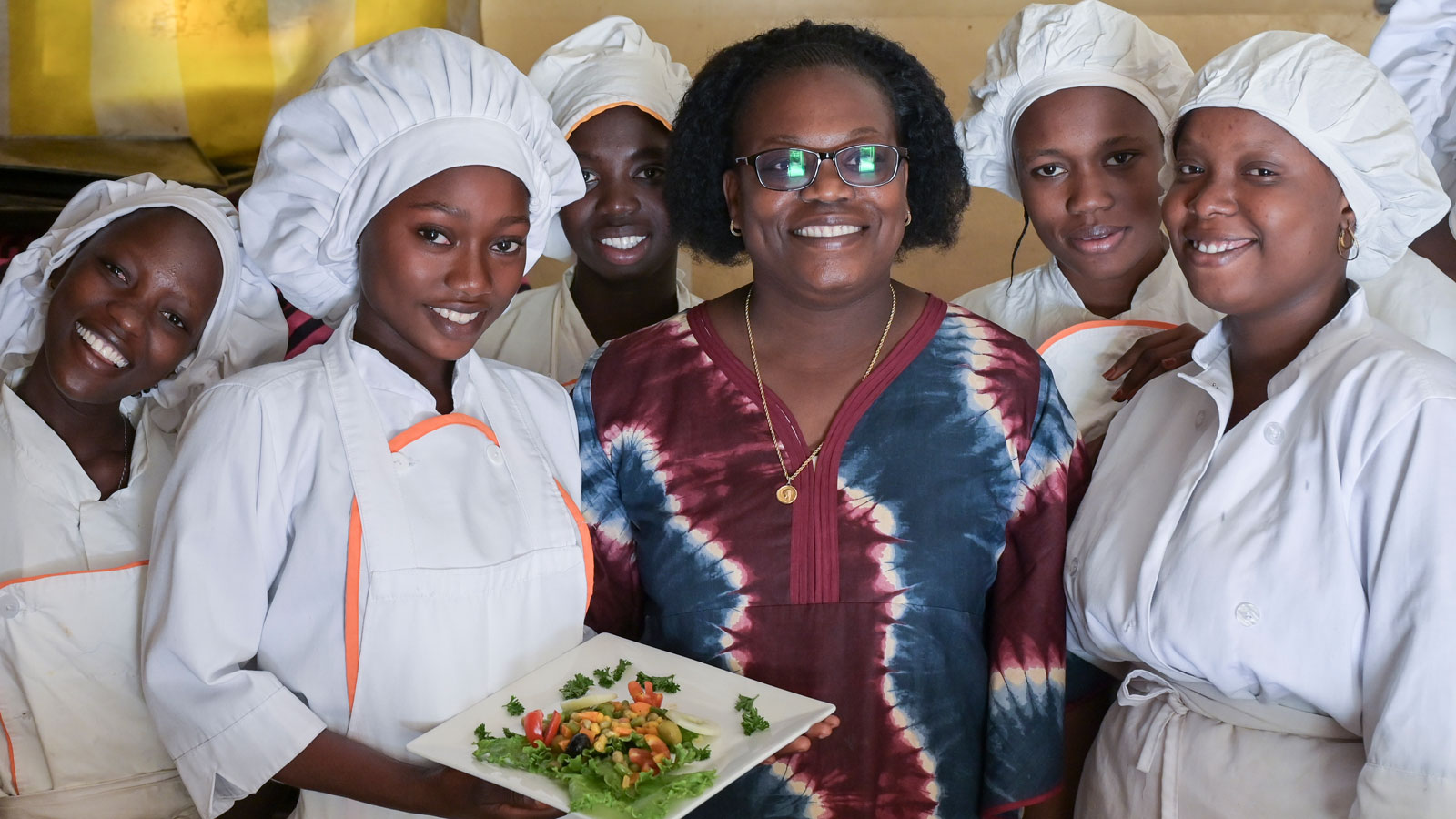 Fünf junge Frauen stehen lächelnd in weißer Küchenkleidung um Louise Ndione, Leiterin der gemeinnützigen Ausbildungseinrichtung Claire Amitié in Thies, Senegal. Die Schülerin Marie steht links von der Leiterin und präsentiert einen Vorspeisensalat.