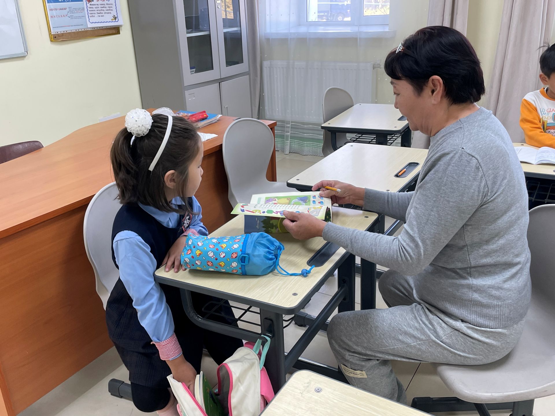 Ein Mädchen packt etwas aus seinem Schulrucksack, während eine Betreuerin ein Buch aufschlägt. Bildunterschrift bei Aktivierung: Die Schülerinnen und Schüler erhalten in der Nachmittagsbetreuung auch Hausaufgabenhilfe.