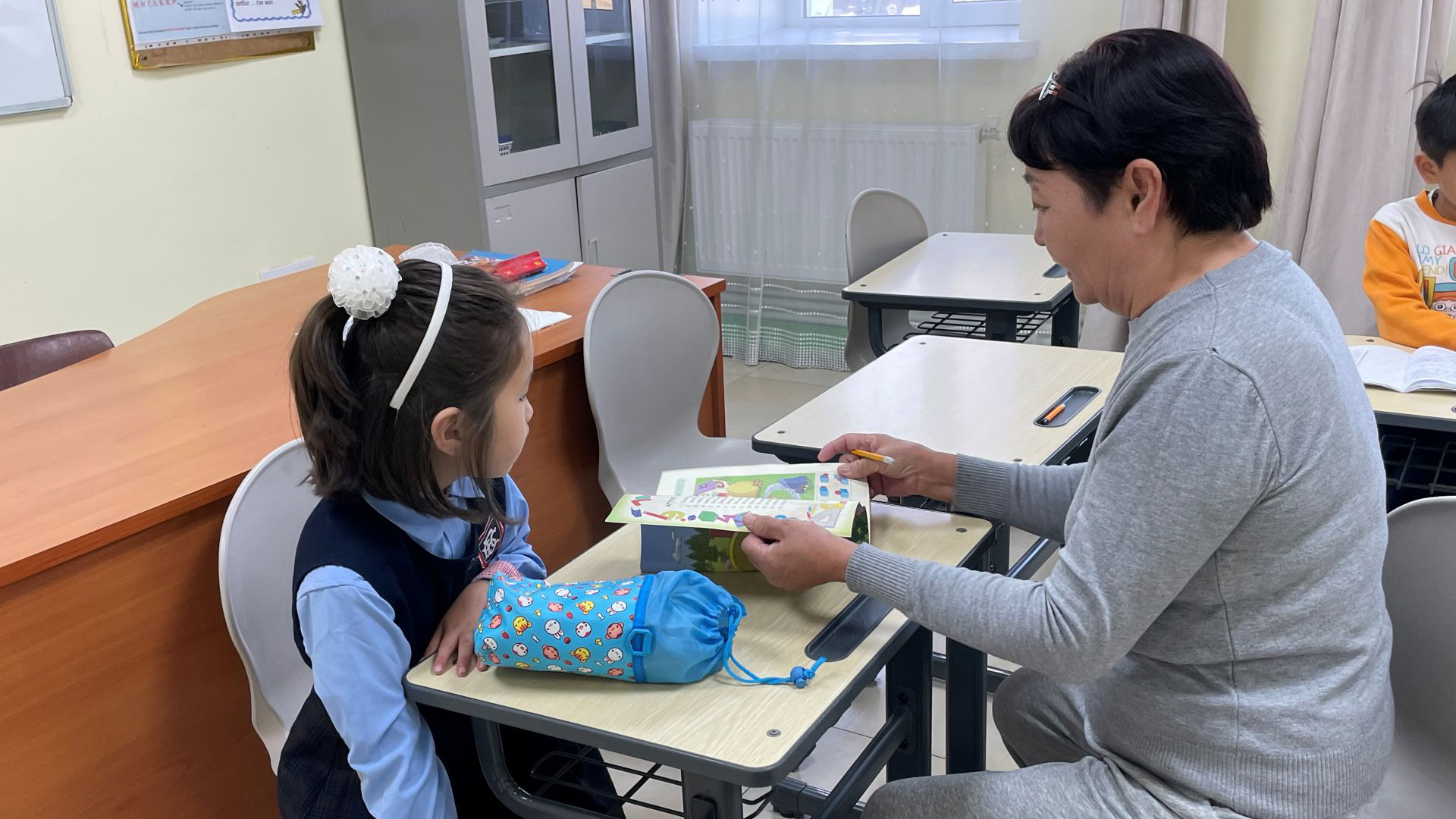 Ulanbaatar, Mongolei: Ein Mädchen packt etwas aus seinem Schulrucksack, während eine Betreuerin ein Buch aufschlägt.