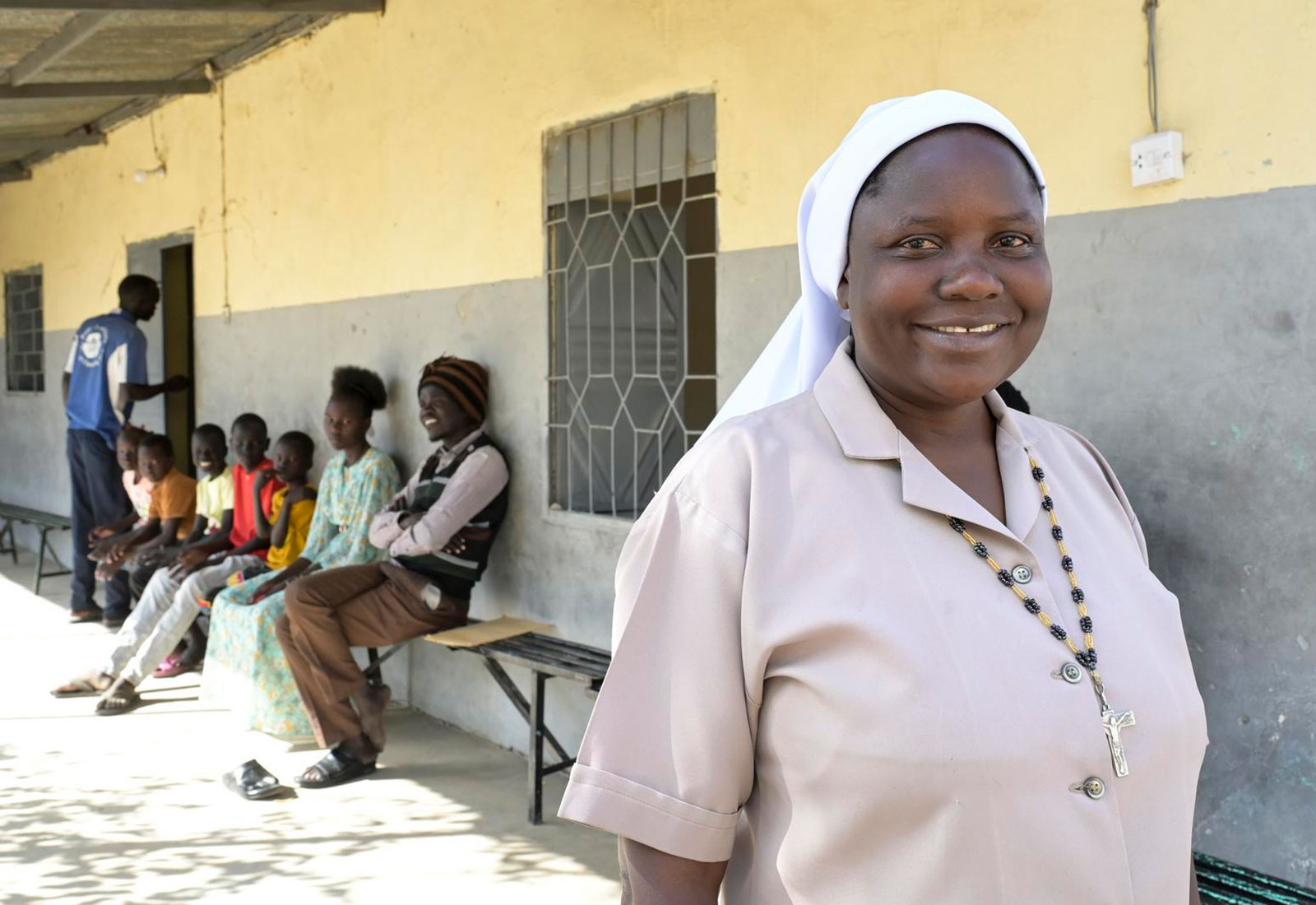 Schwester Amony Susan Atube läuft außen an einem Genbäude vorbei und lächelt in die Kamera. Hinter ihr sitzen auf einer Bank mehrere Kinder und zwei junge Erwachsene.