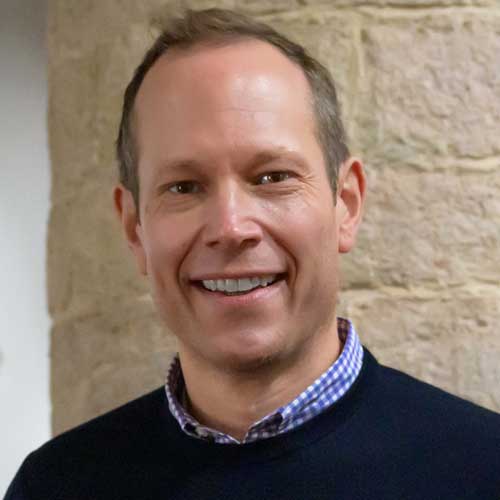 Porträt Christoph Michel. Er trägt ein blau-weiß kariertes Hemd, darüber einen dunklen Pullover und lächelt.