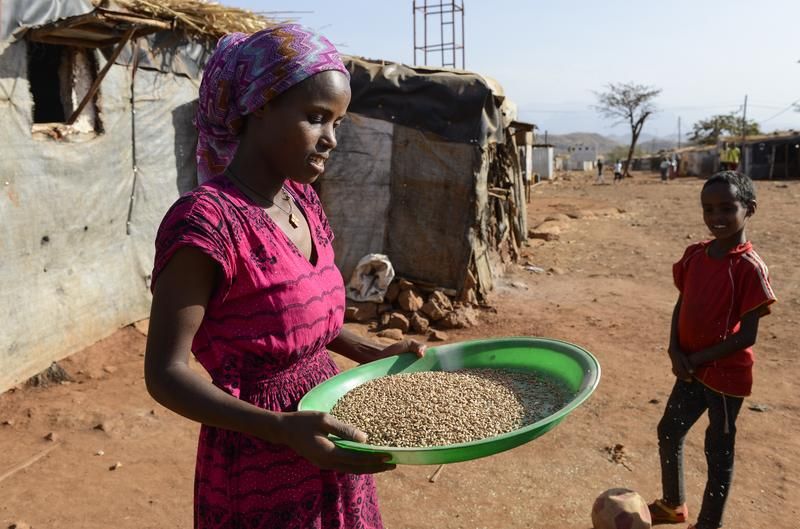 Eine junge Frau steht in Äthiopien am Rande einer Siedlung mit Häusern aus Plastikplanen, Holz und Stroh und hält eine große grüne Plastikschüssel voller Getreide in der Hand. Auf der unasphaliterten Straße steht schräg neben ihr ein kleiner Junge mit einem platten Fußball am Fuß.
