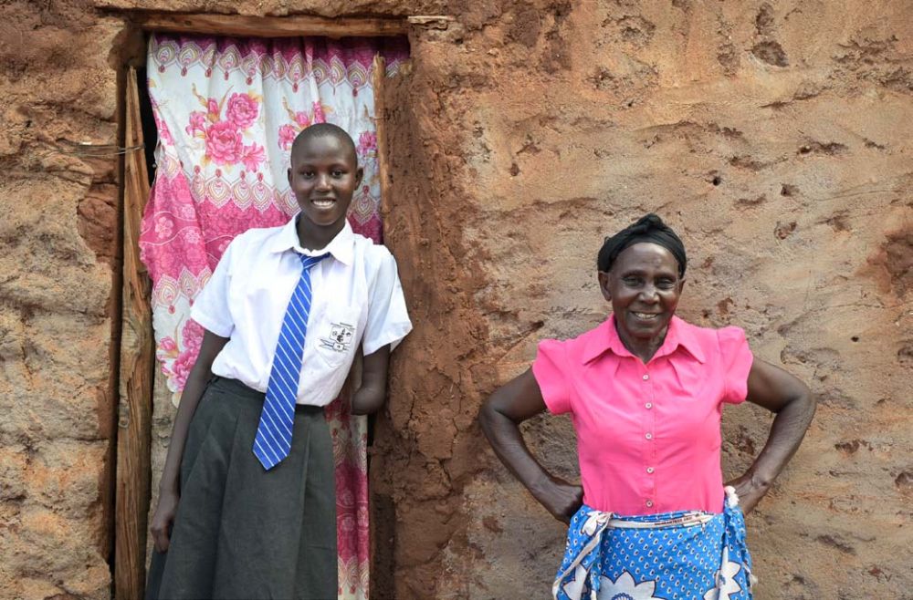 Vor einem Haus aus Lehm mit einer Tür, die allerdings nur aus einem weiß-rosa Vorhang besteht, stehen zwei Frauen: Eine Schülerin in Schuluniform (Rock und weiße Bluse und Krawatte) und daneben eine ältere Frau mit rosa Bluse und blauem Wickeltuch.