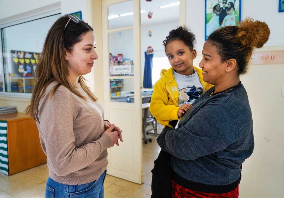 Zwei Frauen unterhalten sich vor einem Klassenzimmer. Die Frau rechts im Bild hat ein kleines Mädchen auf dem Arm, das in die Kamera lächelt.
