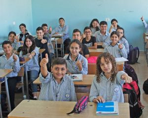 Schulkinder aus dem Libanon sitzen in hellblauer Schuluniform in einem Klassenzimmer an Schultischen. Sie lachen in die Kamera, einige von ihnen strecken ihren Daumen nach oben.