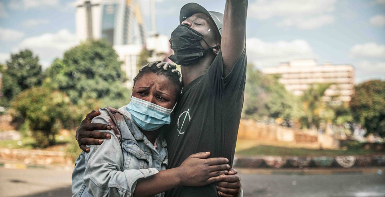 Proteste gegen neues Steuergesetz in Kenia im Juni 2024 – eine Frau mit medizinischer Maske blickt verängstigt und legt die Arme um einen Mann, der die linke Hand nach oben reckt. Die rechte hat er ihr um die Schulter gelegt, die blutverschmiert zu sein scheint.