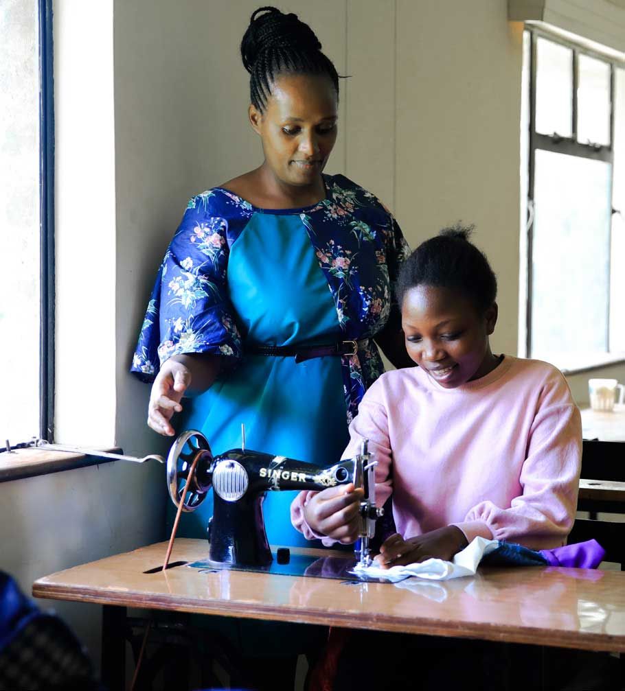 Eine Frau steht in einem Schulungsraum in Kenia hinter einer jungen vor ihr an einer Nähmaschine sitzenden Frau und sieht ihr beim Nähen über die Schultern.
