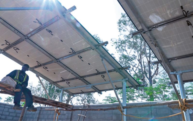 Die Sonne scheint zwischen den zwei Teilen eines Solardachs in Uganda hindurch. Auf dem einfachen Gerüst darunter sitzt ein Arbeiter und macht Pause.