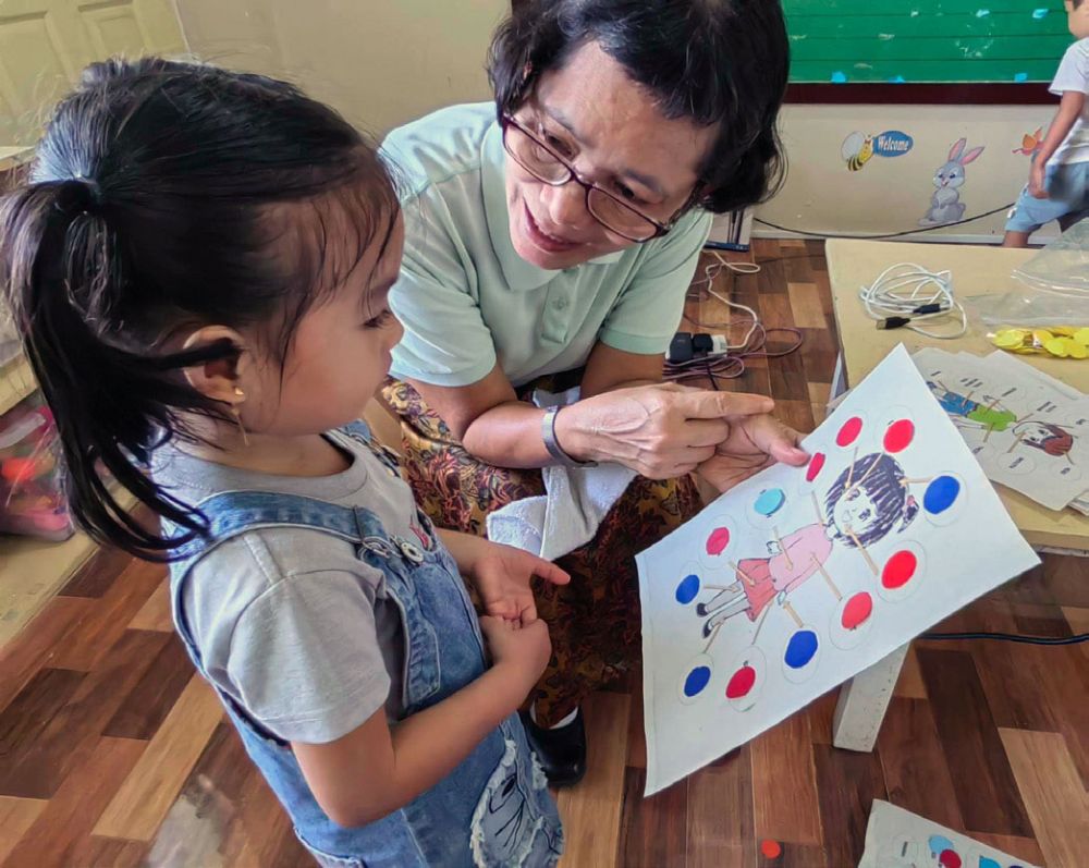 Eine Lehrerin beugt sich zu einer Vorschülerin herunter und zeigt Ihr auf einem Blatt Papier die Zeichnung eines Mädchens.