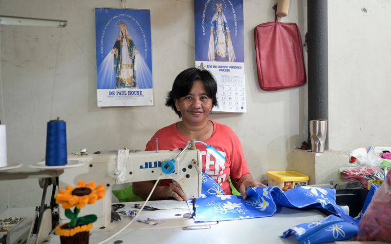 Eine Frau aus den Philippinen sitzt im Armenviertel Payatas in einem Raum an einem Tisch. Vor sich eine Nähmaschine und näht. Hinter ihr an der Wand zwei Kalender mit der heiligen Mutter Maria.