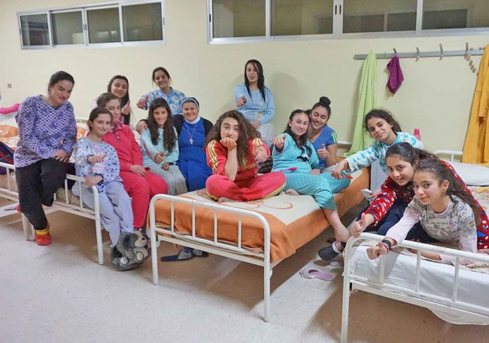 Mädchen und junge Frauen sitzen in einem Schlafsaal des Wohnheims im Libanon auf drei Metallbetten und lachen in die Kamera. Zwischen ihnen sitzt eine Ordensfrau.