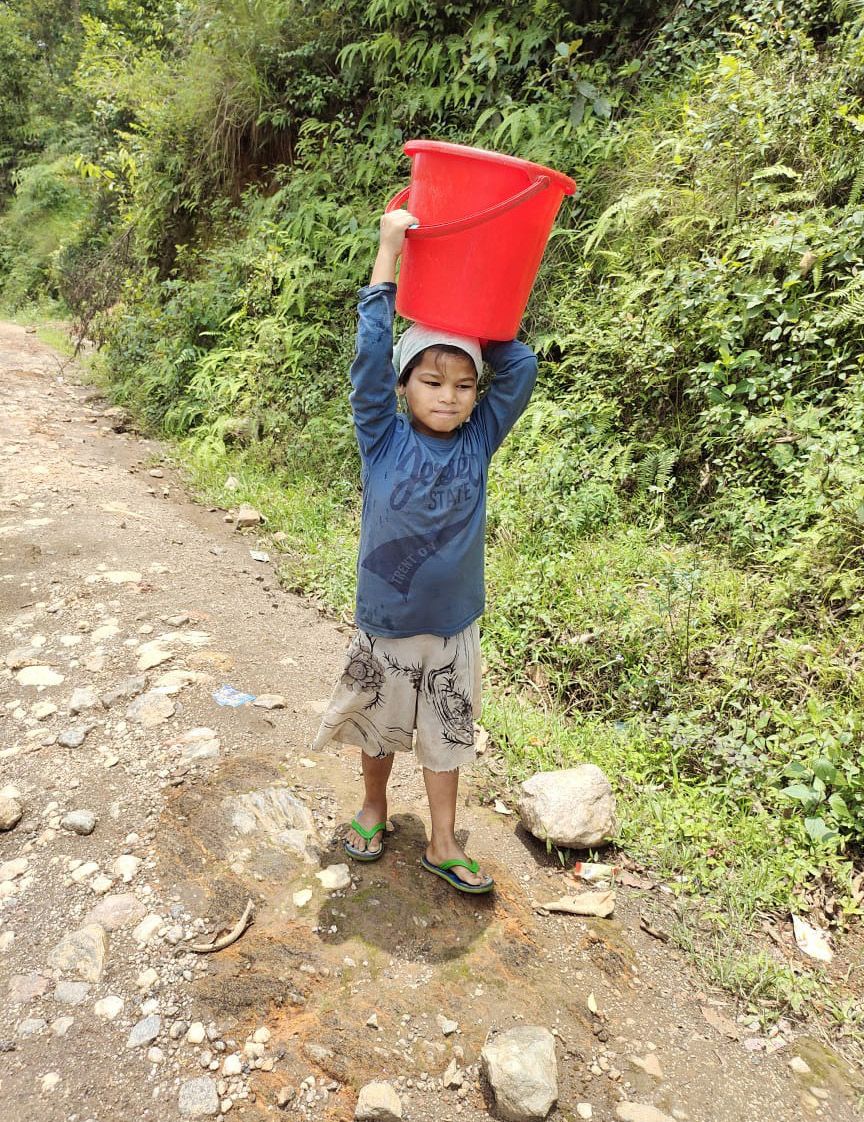 Ein kleiner Junge mit Flip-Flops und kurzen Hosen trägt einen Eimer voller Wasser auf seinem Kopf einen steinigen Weg entlang.