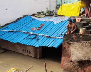 Flut in Indien: Ein Mann sitzt erhöht auf einer Mauer neben einer Wellblechhütte, die zu großen Teilen bereits überschwemmt ist.