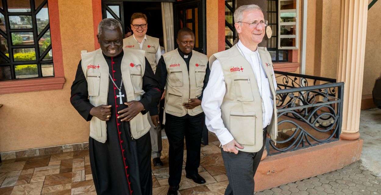 Erzbischof Philip Anyolo (links) und Erzbischof Dr. Stefan Heße (rechts) treten in Githurai im Großraum Nairobi aus einem Gebäude. Sie tragen beige Westen mit missio-Logo. Hinter ihnen folgen weitere Personen.