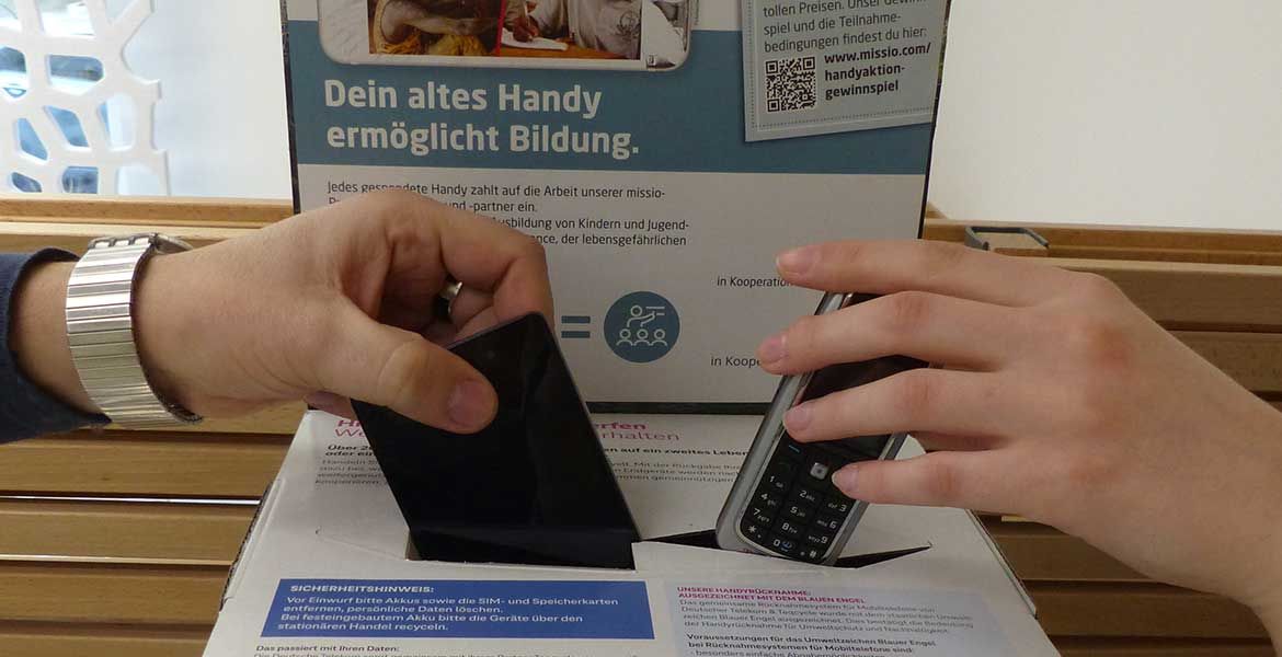 Zwei Hände werfen von links und rechts ausrangierte Handys in eine Spendenbox.