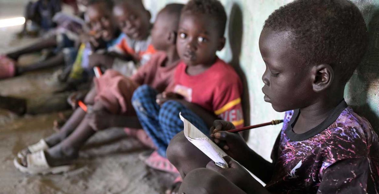 Im Vordergrund rechts im Bild sitzt ein Junge aus dem Südsudan auf dem Boden und schreibt mit einem Bleistift konzentriert in ein Heft. Im Hintergrund neben ihm sind weitere Kinder zu sehen, die zu ihm blicken.
