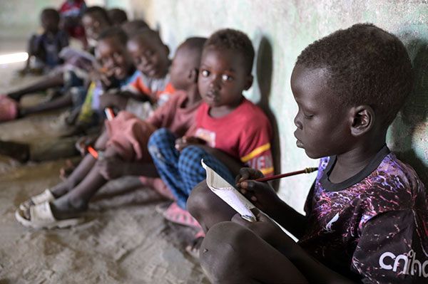 Kinder sitzen an einer Wand. Das erste Kind schreibt kniend etwas in ein Heft.