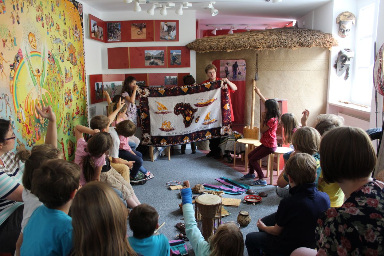 Zu sehen ist der missio-Ausstellungsraum in München. Kinder sitzen auf Hockern im Kreis. Vorne hält eine missio-Mitarbeiterin ein großes Tuch vor sich ausgebreitet in die Luft.