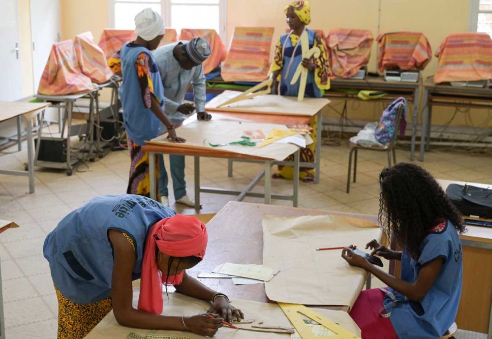 Einblick in die Schneiderwerkstatt des Hilfsprojekts: An zwei großen Tischen stehen vier junge Frauen und ein junger Mann, sie sind über die Tische gebeugt und zeichnen Schnittmuster. Im Hintergrund stehen aufgereiht zehn mit Stoffen zugedeckte Computer. 