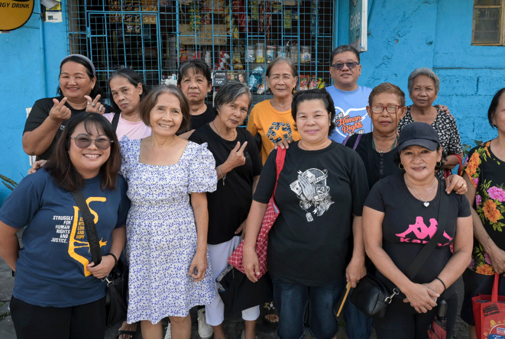 Father Toots steht mit einer Gruppe von Frauen, darunter eine Mitarbeiterin von TFDP, vor einem geschlossenen kleinen Geschäft. Foto: Jörg Böthling