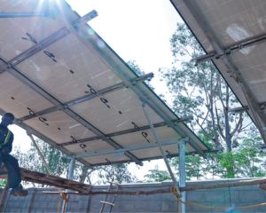 Die Sonne scheint zwischen den zwei Teilen eines Solardachs in Uganda hindurch. Auf dem einfachen Gerüst darunter sitzt ein Arbeiter und macht Pause.