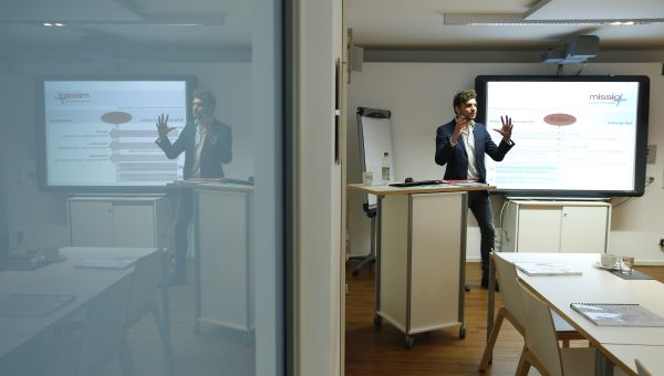 Zu sehen ist ein Seminarraum mit einem Referenten an einem Stehpult. Hinter ihm ein Monitor.