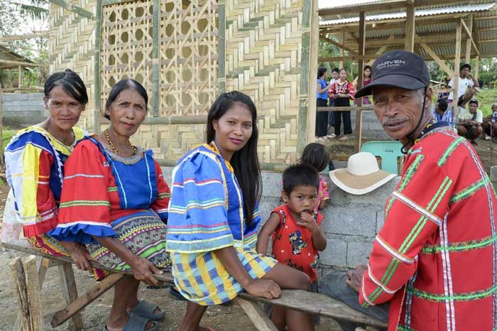 Drei Frauen und ein Mann aus der indigenen Bevölkerungsgruppe in Mindanau auf den Pilippinen sitzen in bunten, folkloristischen Kleidern auf einer Holzbank vor einem Gebäude aus Bambus. Ein kleines Kind steht neben einer der sitzenden Frauen und sieht ebenfalls in die Kamera.