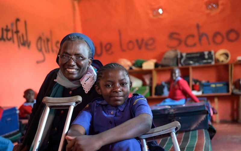 In einem Klassenzimmer mit rot-orangenen Wänden ist im Vordergrund eine Ordensschwester aus Kenia zu sehen, die neben einem Mädchen mit Behinderung sitzt. Vor der Ordensschwester lehnt die Krücke des Mädchens.