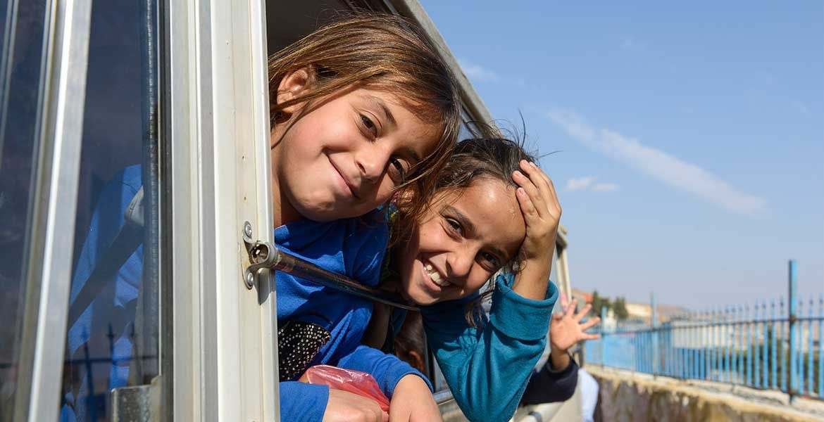 Zwei Mädchen schauen aus einem Busfenster und lachen in die Kamera.
