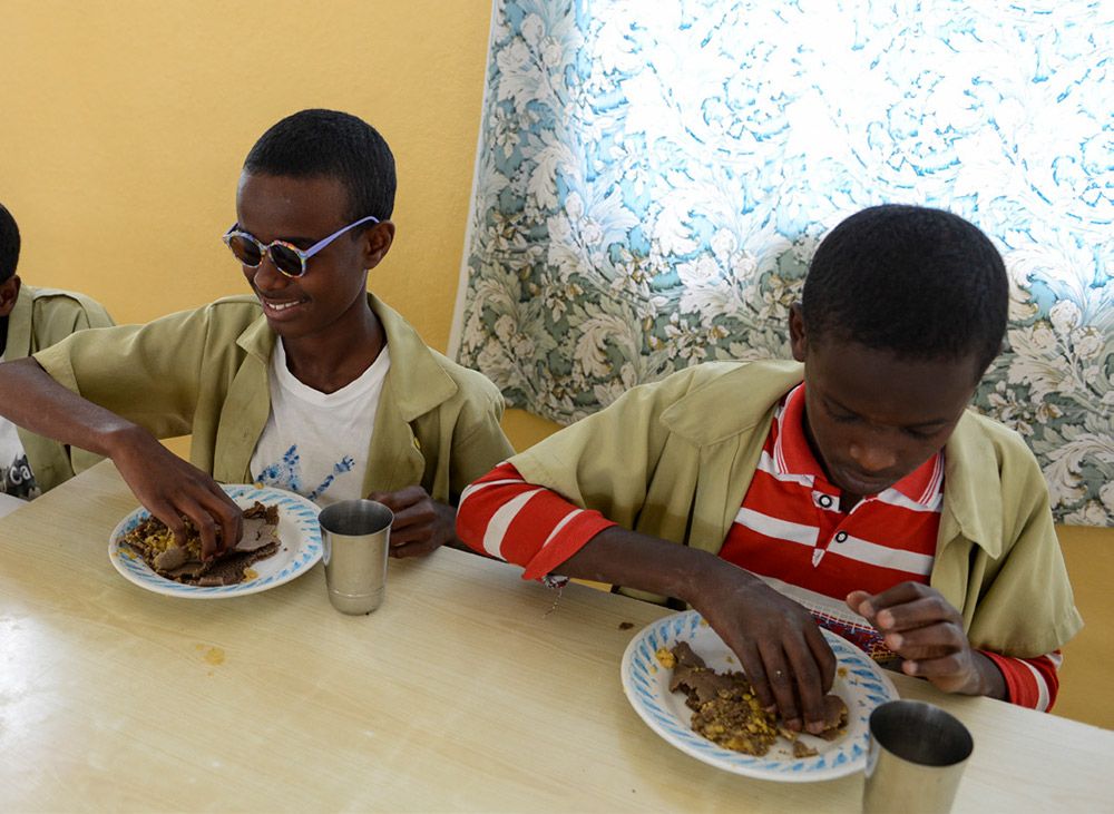 Zwei Schüler der Blindenschule sitzen an einem Esstisch und essen mit den Händen ihr Mittagessen.