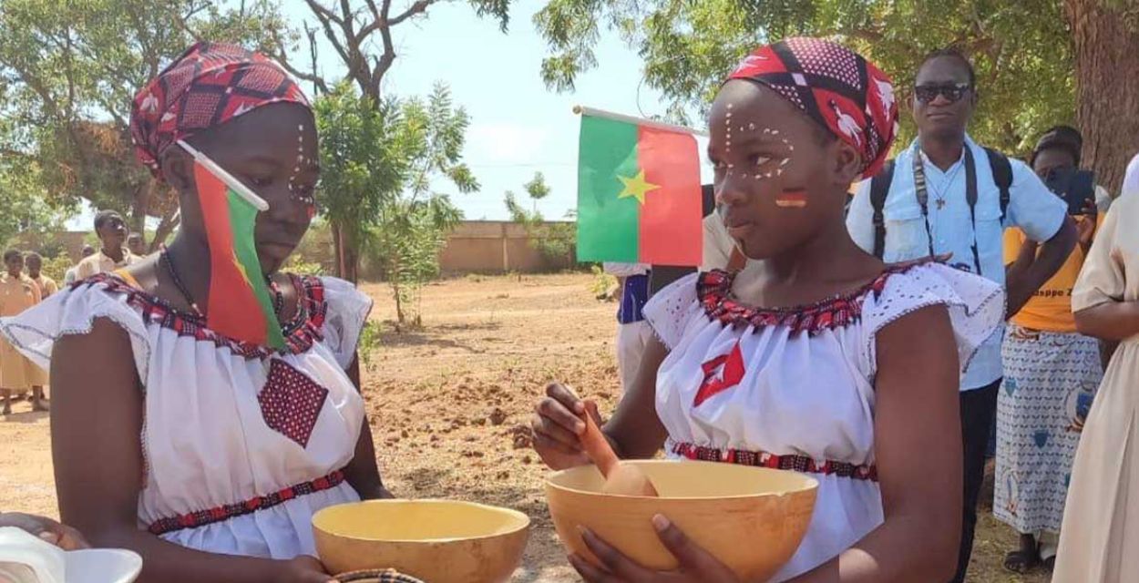 Zwei junge Frauen aus Burkina Faso haben zwei Holzschüsseln in der Hand und sind geschmückt mit burkinischen und deutschen Flaggen.